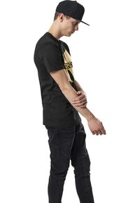 Camiseta negra con un gran logo amarillo en el frente, combinada con jeans oscuros que tienen acentos texturizados. Modelo llevando una gorra negra.