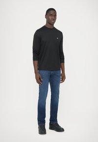 Must pika varrukaga särk ümmarguse kaelusega, koos siniste sirge säärega teksade ja mustade tossudega, seisab neutraalsel taustal.