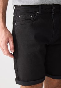 Crne traperice kratke hlače s glatkom teksturom, s preklopljenim rubovima, metalnim dugmetima i prednjim džepovima. Vidljivo je detaljno šivanje.