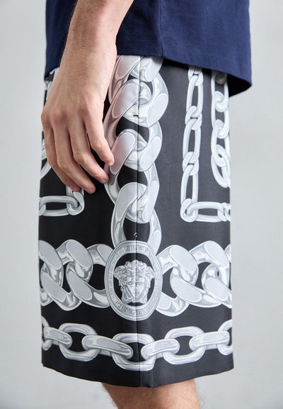 Shorts noirs avec un motif de chaîne blanche et un médaillon circulaire représentant un visage. Les shorts ont une texture lisse et une coupe ajustée.
