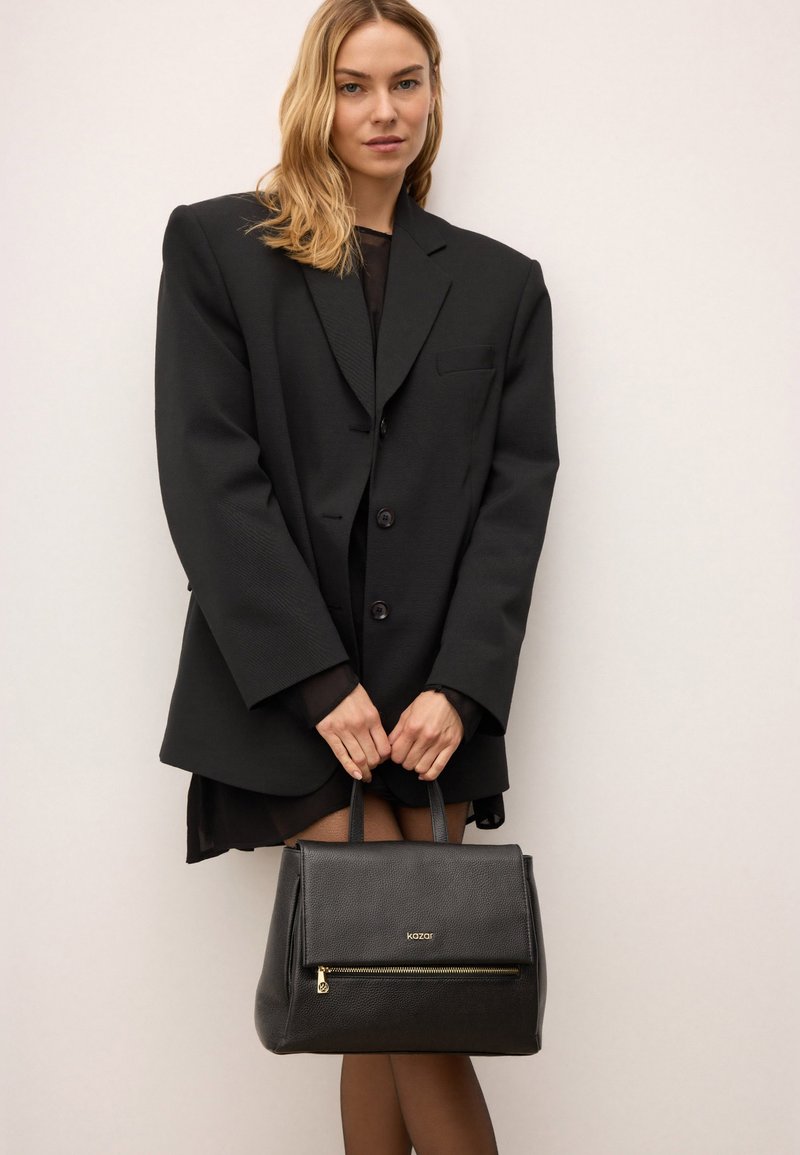 Borsa a mano in pelle nera con forma strutturata, dotata di chiusura zip superiore e dettagli in metallo dorato, tenuta da una persona che indossa un blazer oversize.