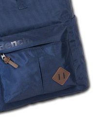 Marineblauwe rugzak van duurzaam materiaal, met een voorvak met rits, bruine leren accenten en een diamanten patch met uitsnijdingen.