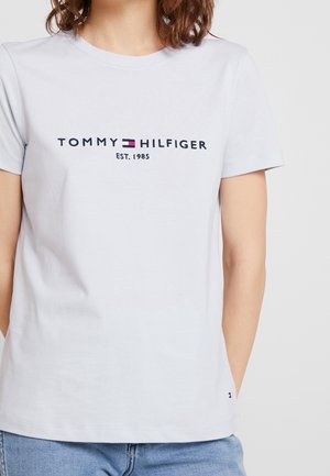 Biała koszulka z krótkim rękawem z "Tommy Hilfiger Est. 1985" i logo marki noszona przez osobę z rękami w kieszeniach.
