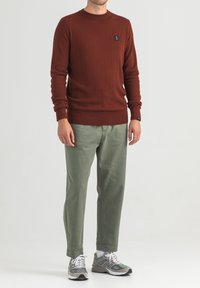 Maglione a maglia croccante di colore marrone con collo a girocollo, abbinato a pantaloni verde chiaro. Il modello indossa scarpe da ginnastica grigie. Logo nero sul davanti del maglione.