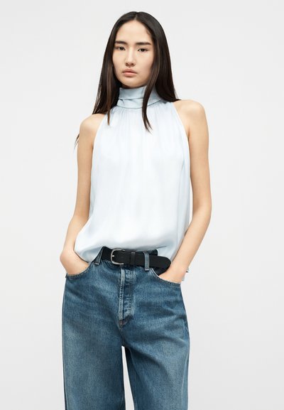 Ung kvinne med langt mørkt hår iført en ermeløs hvit bluse med høy hals, stappet inn i løstsittende blå jeans med svart belte, hendene i lommene.