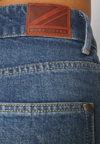 Plave traperice od denima s smešnim kožnim etiketom koja ima crvene pruge i urezana slova "PEPE JEANS". Uključuje žute šavove i metalne detalje.