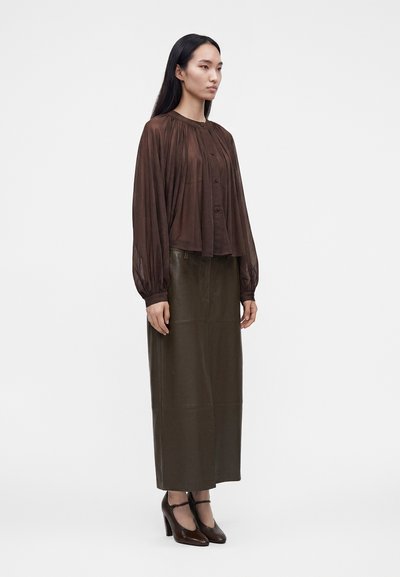 Femme debout portant un chemisier marron transparent à manches longues, une longue jupe vert olive et des chaussures marron à talons hauts avec brides à la cheville.