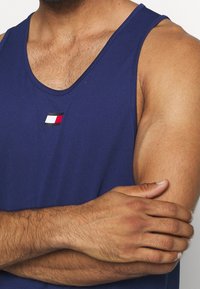 Uomo con un canottiera blu senza maniche e un piccolo logo rettangolare rosso, bianco e nero sul petto, braccia incrociate sul torso.