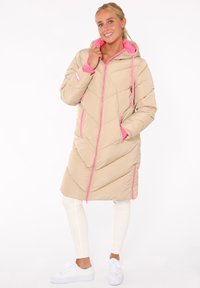 Beige Pufferjacke mit rosa Akzenten, gesteppter Optik, Kapuze und Reißverschluss. Verfügt über zwei Seitentaschen und einen mittellangen Schnitt.