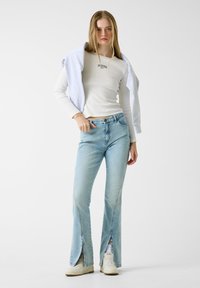 Ανοιχτό μπλε τζιν καμπάνα με πλαϊνές σχισμές, συνδυασμένο με εφαρμοστή μακρυμάνικη μπλούζα εκρού. Μια ανοιχτό γκρι φούτερ φαρδιά φορεμένη πάνω από τους ώμους.