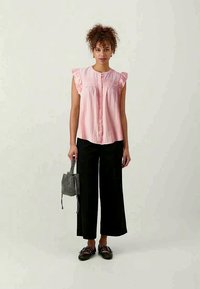 Ροζ αμάνικο μπλουζάκι με κουμπιά μπροστά, φρουτιέρες λεπτομέρειες και υφασμάτινα στοιχεία, συνδυασμένο με μαύρα παντελόνια με φαρδιά πόδια και μια μικρή τσάντα με σχέδιο.