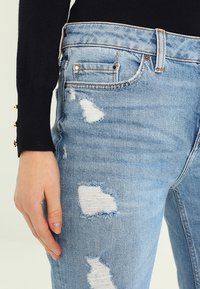 Jeans in denim blu chiaro invecchiato presentano accenti strappati e un classico design a cinque tasche. Il materiale mostra leggeri segni di usura e una texture morbida.