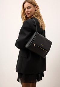 Borsa a spalla in pelle nera con design a patta, caratterizzata da una cerniera dorata e dettagli con logo. Texture liscia con una forma strutturata.