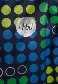 Námorní modrá látka s velkými zelenými a modrými kruhovými vzory. Ve středu je umístěno stříbrné kruhové logo s nápisem "LEGO", obklopené menšími kruhy.