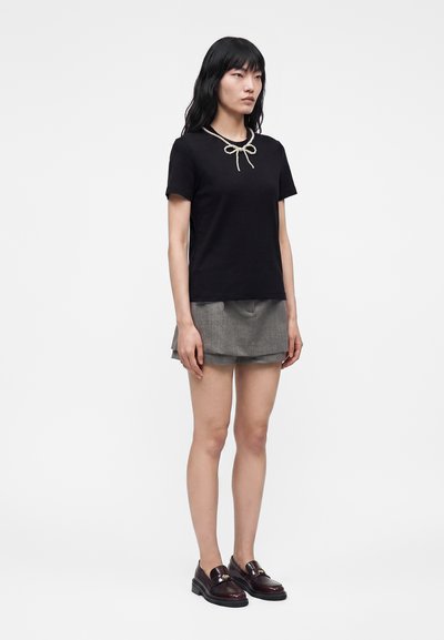 Femme aux longs cheveux foncés portant un T-shirt noir avec un collier nœud en perles, une mini-jupe grise à plusieurs couches et des mocassins foncés, debout sur fond blanc.