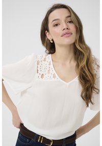 Blouse blanche en tissu texturé, avec un détail en dentelle à l'encolure, décolleté en V et manches courtes. Portée avec une ceinture marron et un jean noir.
