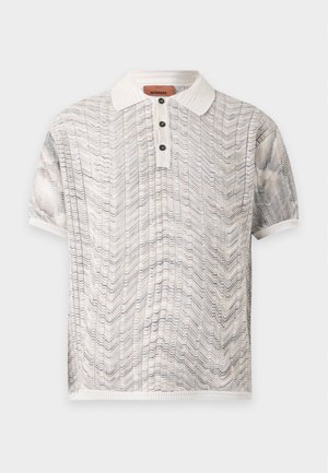 Jeune homme portant des lunettes de soleil foncées, un polo en maille beige à manches courtes, un jean large bleu clair et des chaussures marron foncé, debout sur fond blanc.
