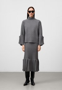 Maglione grigio a maglia con collo alto e maniche a campana, abbinato a una gonna coordinata con pieghe sovrapposte. La modella indossa occhiali da sole neri e stivali.