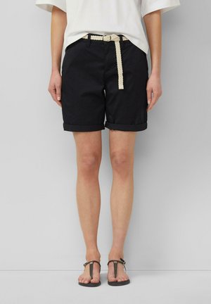 Staand persoon, gekleed in zwarte opgerolde shorts met een witte geweven riem, wit T-shirt en zwarte teenslippers tegen een effen achtergrond.