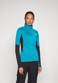 Žalias zip-up tipo džemperis su tekstūruotu korpusu, juodomis rankovėmis, šoninėmis kišenėmis ir aukštu kaklu. Ant krūtinės yra logotipas ir kontrastingi siūlai.