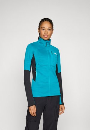 Teal zip-up jakna z teksturiranim telesom, črnimi rokavi, stranskimi žepi in visokim ovratnikom. Ima logo na prsih in kontrastne šive.