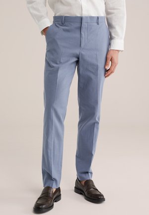 Mand iført lyseblå skræddersyede bukser, hvid skjorte og mørkebrune læder loafers, stående mod en ensartet baggrund.