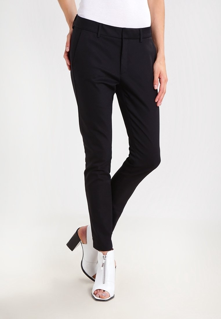 Pantalon noir slim porté par une personne avec des bottines blanches à bout ouvert et talon, debout jambes croisées sur un fond uni.