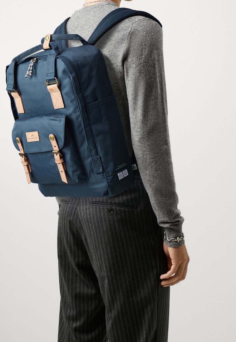 Personne portant un sac à dos bleu marine avec des bretelles en cuir beige, un pull gris et un pantalon noir à fines rayures, vue de dos et de profil.