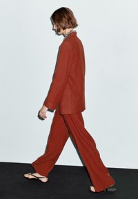 Completo di lino color ruggine con blazer strutturato e pantaloni a gamba larga, abbinato a infradito nere, su uno sfondo neutro.