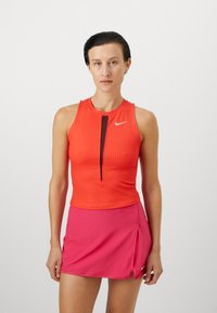 Ärmelloses Sportoberteil in leuchtendem Rot mit gerippter Textur und einem zentralen schwarzen Reißverschluss. Kombiniert mit einem pinken Sportrock mit seitlicher Schlitze.