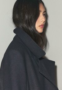 Sort uldfrakke med høj krave og struktureret silhuet. Glat tekstur; synlige stikningsdetaljer langs kanterne. Neutral baggrund.