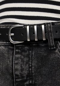 Cinto de couro preto com fivela de metal prateada com três garras, usado sobre uma blusa preta riscada e tecido de denim escuro.