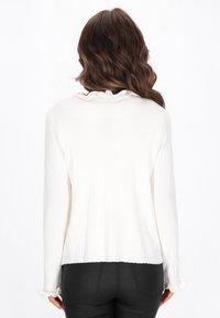 Fehér bordás, hosszú ujjú felső fodros nyakkivágással és mandzsetta részletekkel, laza szabású és sima textúrával.