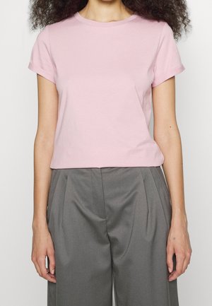 Vrouw draagt een lichtroze T-shirt met korte mouwen, ingestopt in een losse, hooggetailleerde grijze broek met plooien aan de voorkant.