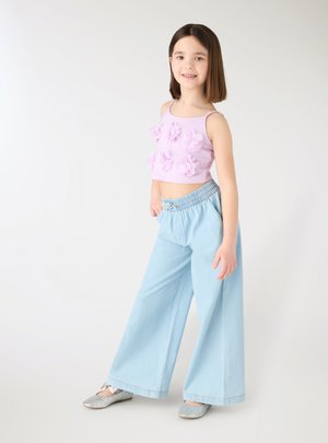 Ragazza giovane che sorride, indossa una canotta viola chiaro con fiori di tessuto, pantaloni a gamba larga azzurri e ballerine argento.