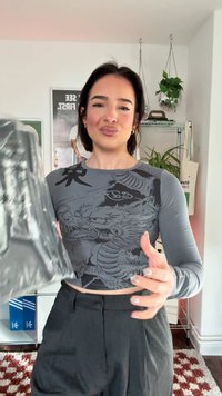 Grijze crop top met lange mouwen, met een gedetailleerde zwarte draakprint en tekst. Model draagt een glanzende zwarte tas, met donkere broeken en een gestructureerde achtergrond.