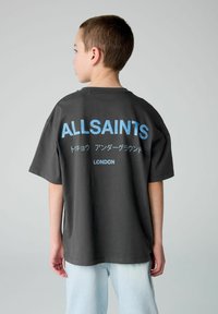 Pilkos spalvos per didelis marškinėliai su trumpomis rankovėmis, ant nugaros su mėlynu tekstu ir simboliais, įskaitant "ALLSAINTS" ir "LONDON."