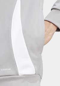 Graue Sportjacke mit weißem Seitenpanel, aus texturiertem Stoff, mit fetten Ärmeln und "AEROREADY"-Logo in Weiß auf der unteren linken Seite.