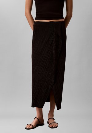 Γυναίκα με μαύρο αμάνικο τοπ, μαύρη midi φούστα τύπου wrap με υφή υφάσματος και μαύρα φλατ σανδάλια σε απλό γκρι φόντο.