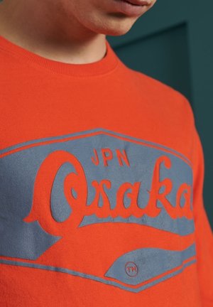 Roter Baumwoll-Sweatshirt mit einem blauen Osaka-Graphic auf der Vorderseite, mit geschwungenem Text und einer glatten Textur.
