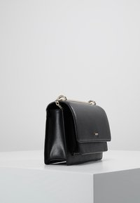 Černá kožená kabelka se zlatým řetízkovým popruhem a klopovým zapínáním, vystavená na bílém povrchu před šedým pozadím.