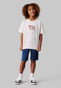 Balta T-krekls ar daudzkrāsainu grafisko dizainu priekšpusē, kopā ar zilām džinsa šortiem. Apģērbu papildina sporta apavi un baltas zeķes.