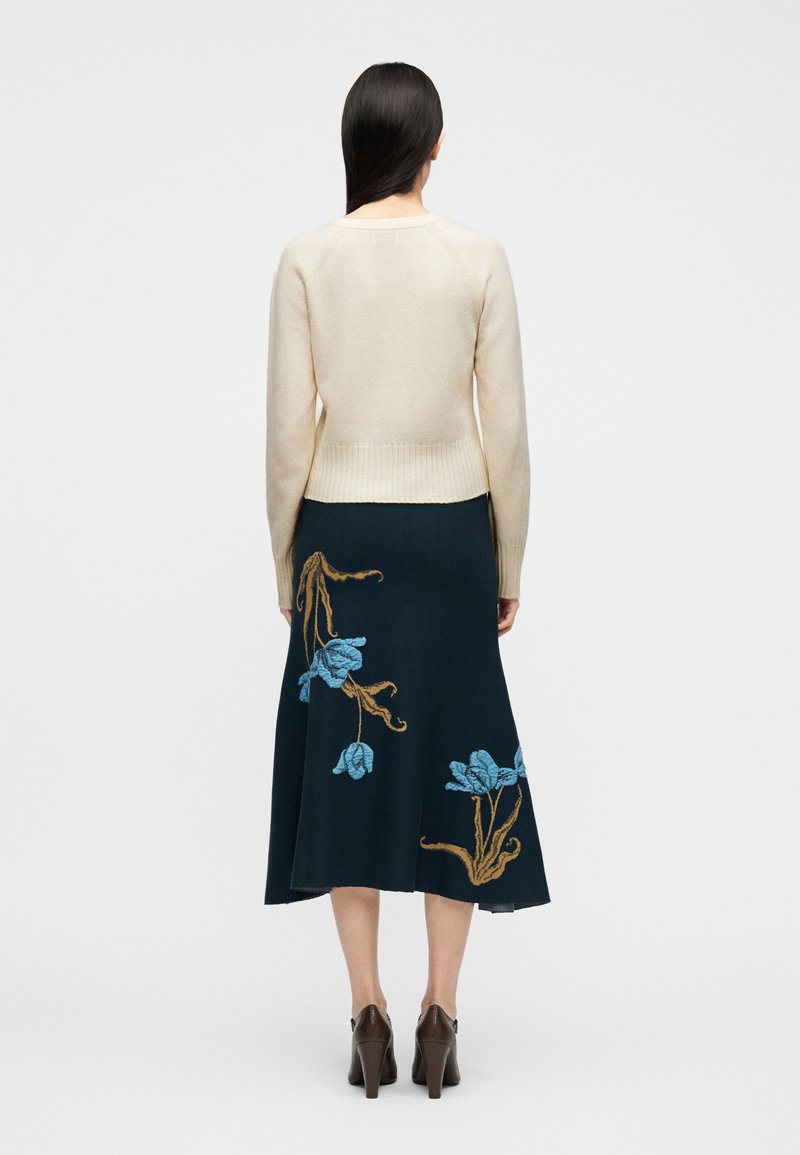 Vrouw met lang donker haar, gekleed in een crème trui en een marineblauwe midi-rok met blauw en bruin bloemenborduursel, gecombineerd met bruine hakken.