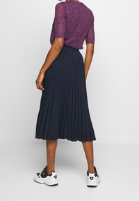 Gonna midi plissettata blu navy abbinata a un top viola luccicante, completata da sneakers bianche e nere, che mette in risalto un fit rilassato e un design fluido.