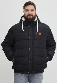 Sort pufferjakke med hætte, forsynet med en faux fur-foring, vandret syning, frontknapper og en logo-patch på brystet.