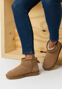 Stivaletti alla caviglia in suede tan con rivestimento interno in pelliccia sintetica, punta rotonda e cuciture decorative, abbinati a jeans stretti blu scuro su uno sfondo di legno.