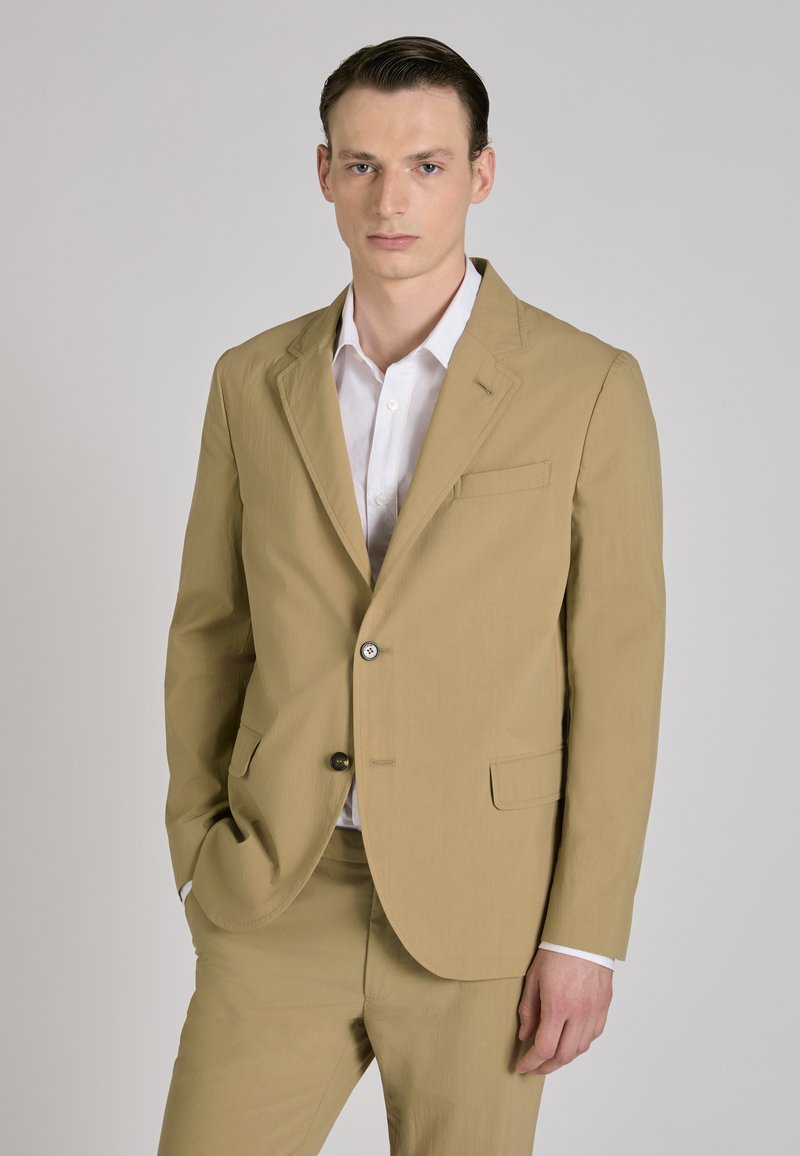 Man met naar achteren gekamd haar draagt een beige pak en wit overhemd, staand met één hand in de zak tegen een effen grijze achtergrond.