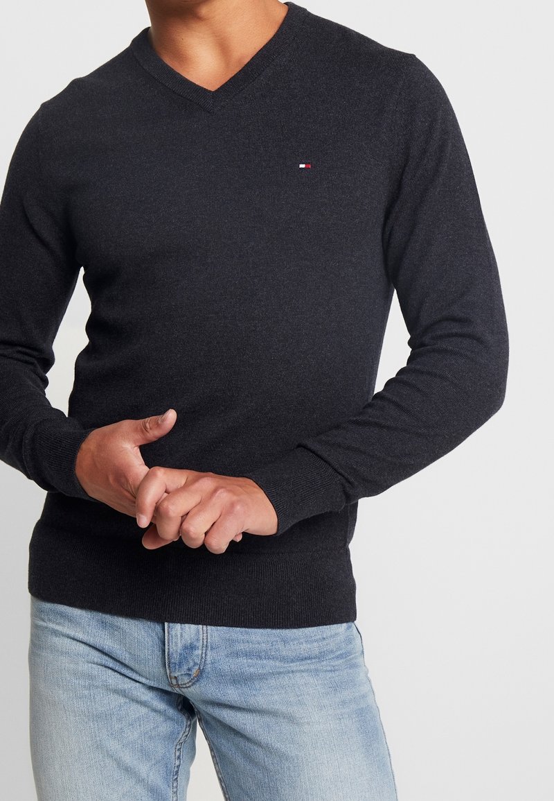 Homme portant un pull noir à col en V avec un petit logo sur la poitrine et un jean bleu clair, debout devant un fond uni.