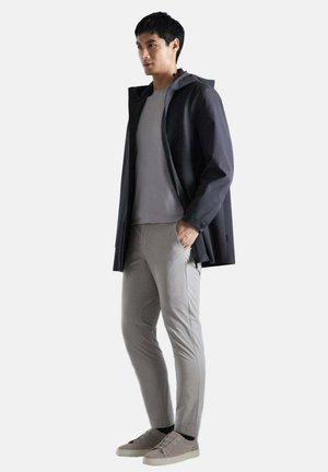 Hombre que lleva una chaqueta con capucha de color oscuro sobre una camiseta gris claro, pantalones ajustados gris claro y zapatillas slip-on gris claro, de pie con una mano en el bolsillo.