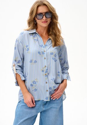 Kobieta o falowanych blond włosach, w dużych ciemnych okularach przeciwsłonecznych, w niebieskiej koszuli z bottonami w paski i kwiatowy wzór oraz jasnoniebieskich dżinsach, pozująca z rękami przy talii.